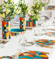 Vista Alegre | Futurismo Bread And Butter Plate on a set table with geometric patterned dinnerware, gold-tone flatware, and floral runner of lilies.