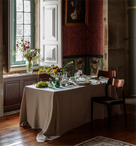 Dining corner featuring Vista Alegre | The Meaning XL Plate on a linen-draped table with fruit bowl, green glass vessels, and elegant tableware, set against historic decor.