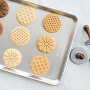 Nordic Ware Heirloom Cookie Stamps on baking sheet, showcasing flower, scallop, and diamond patterns beside a wooden handle, perfect for creating detailed cookie designs.