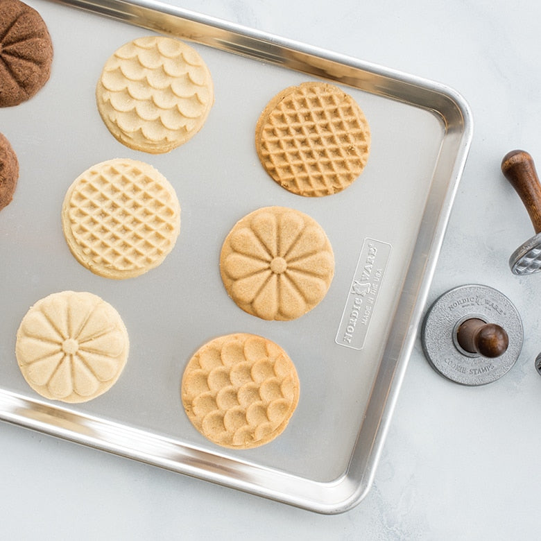 Nordic Ware Heirloom Cookie Stamps on baking sheet, showcasing flower, scallop, and diamond patterns beside a wooden handle, perfect for creating detailed cookie designs.