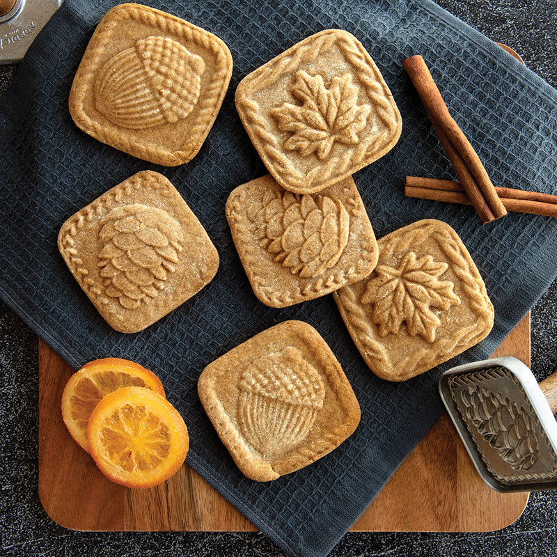 Nordic Ware Fall Forest Springerle Cookie Stamps beside cookies and oranges, showcasing intricate autumn-themed designs for enhancing your seasonal baking.