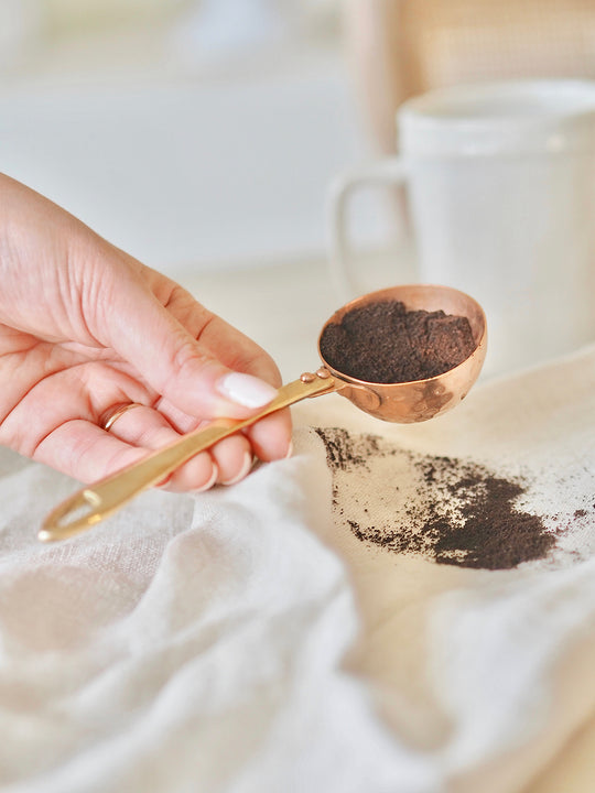Be Home | Camden Scoop: A hand elegantly holds a handcrafted, hammered copper measuring spoon filled with ground coffee, showcasing rustic design by Athens Cooks.