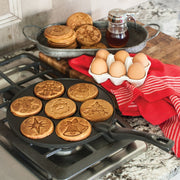 Nordic Ware | Holiday Pancake Pan with festive designs for making seven 3-inch pancakes, shown with pancakes on a pan next to eggs and cookies.