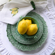 Arte Italica & Crown Linen Designs Renaissance Dinner Plate in green, featuring ornate patterns, topped with lemons and a matching bowl, on a white ruffled placemat.
