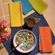 Atelier Saucier | The Toucan Napkins Set of 4 displayed beside a bowl of cereal and a coffee mug on a table.