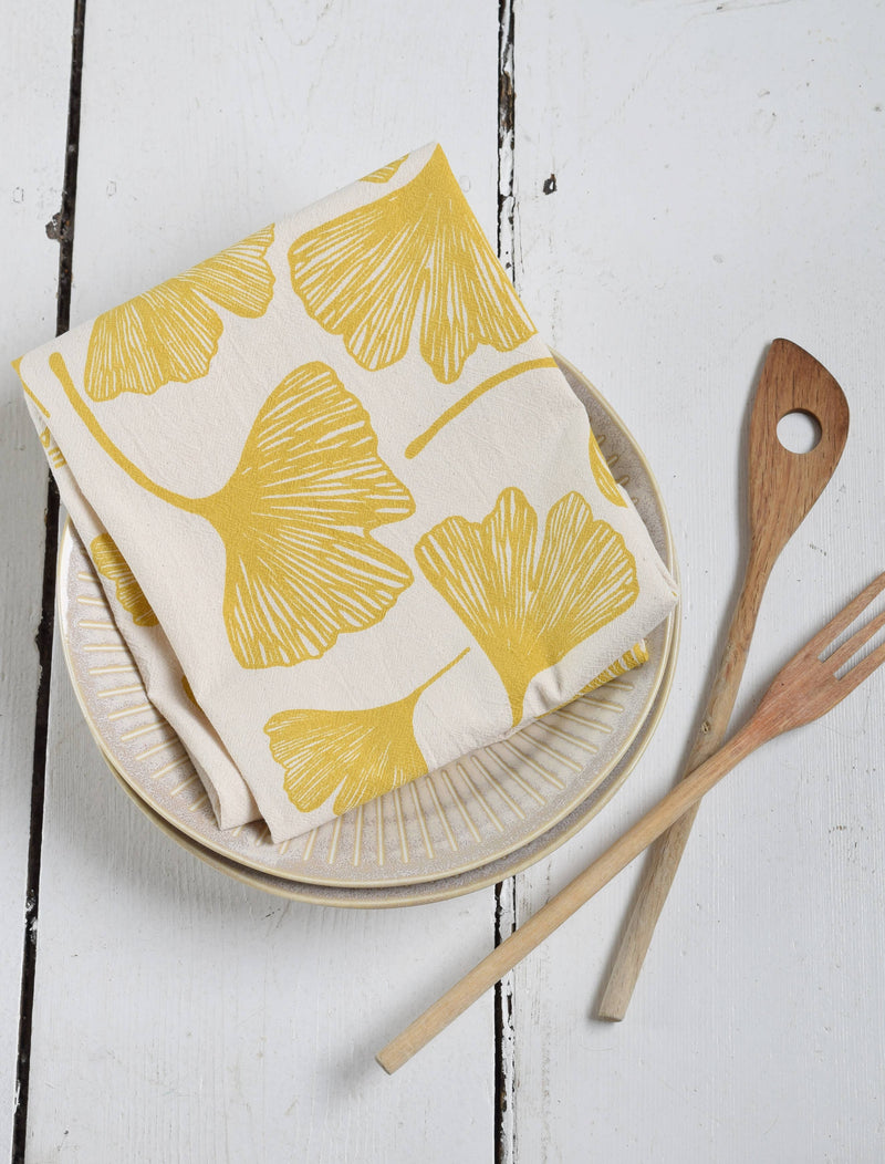 Hearth and Harrow - Ginkgo Leaf Tea Towel - Organic Cotton, displayed folded on ceramic plates with wooden utensils, showcasing its eco-friendly design and oversized 30 x 30 size.