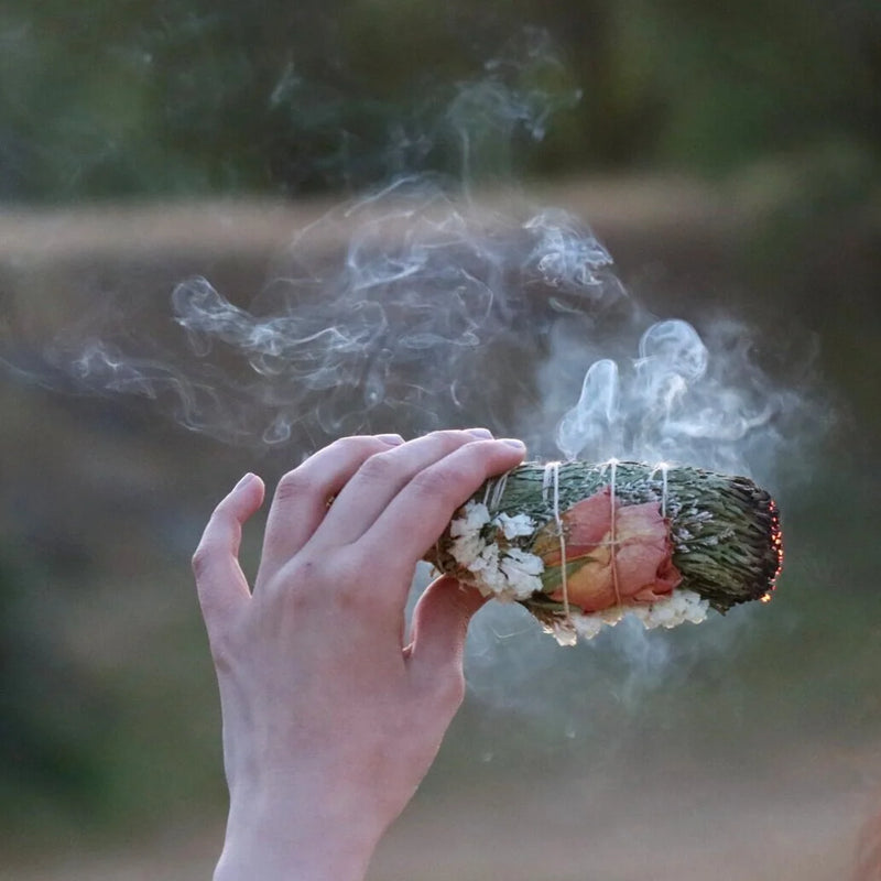 Hand holding Among the Flowers | Floral Smudge Wands, a smoldering bundle of white sage and rose petals, releasing smoke to cleanse and uplift.