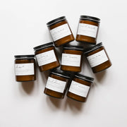 Among the Flowers | Essential Oil Candle, Through the Woods: Eight amber-glass jars with black lids, featuring scented soy candles like “chai” and “ember,” arranged in a circle.