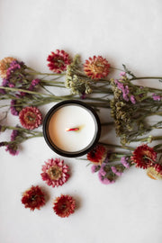 Among the Flowers | Essential Oil Candle, Through the Woods, in a black tin with a wood wick, surrounded by dried flowers on a white surface.