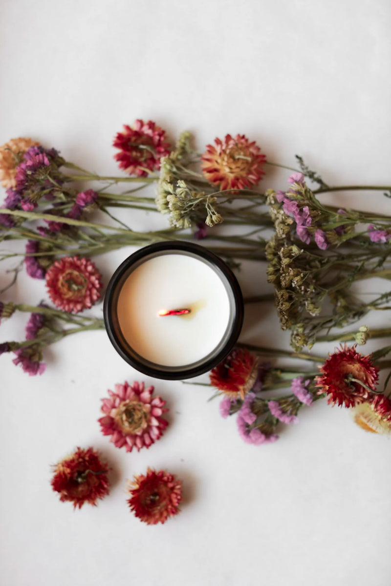 Among the Flowers | Essential Oil + Soy Wax Candle Wilder in a dark jar, surrounded by dried flowers; top-down view showing a slightly singed wick.