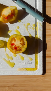 Material - The MK Free Board: Cloud on wooden surface, showcasing a cut yellow tomato and blue-handled peeler, highlighting its non-porous, plant-based design.