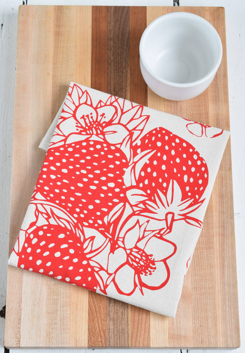 Hearth and Harrow - Strawberry Tea Towel, organic cotton, features a red fruit print design, displayed folded on a wooden surface with a white cup nearby.