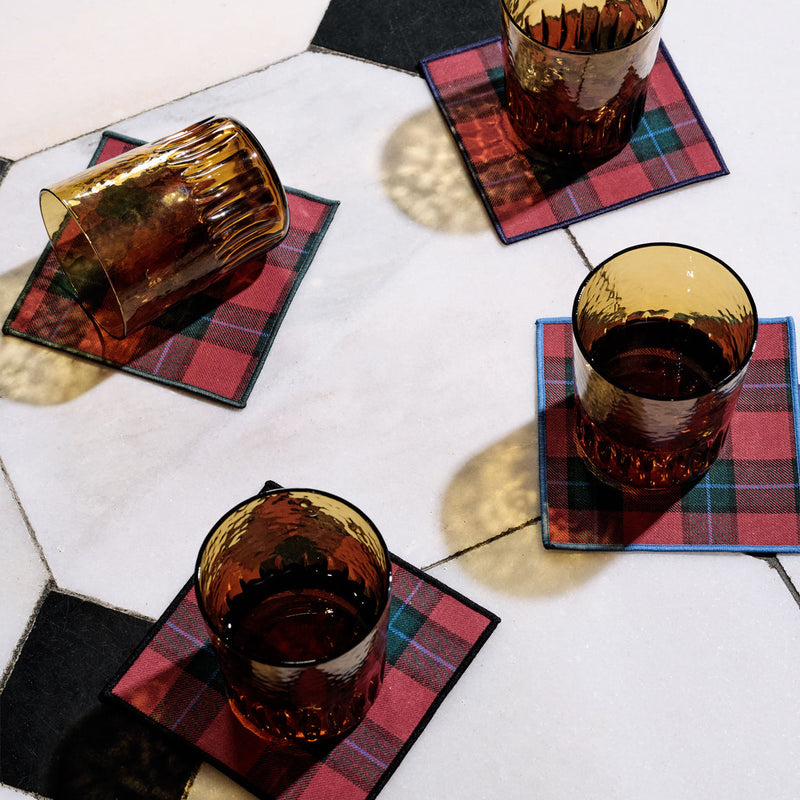 Four amber-tinted glasses on Jubilee Plaid Cocktail Napkins by Atelier Saucier, set on a hexagonal-tiled surface, showcasing festive barware essentials.