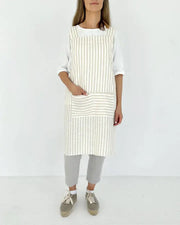 A woman wearing the MagicLinen Pinafore cross-back linen apron, featuring stripes and twin-sectioned pockets, ideal for cooking or gardening.