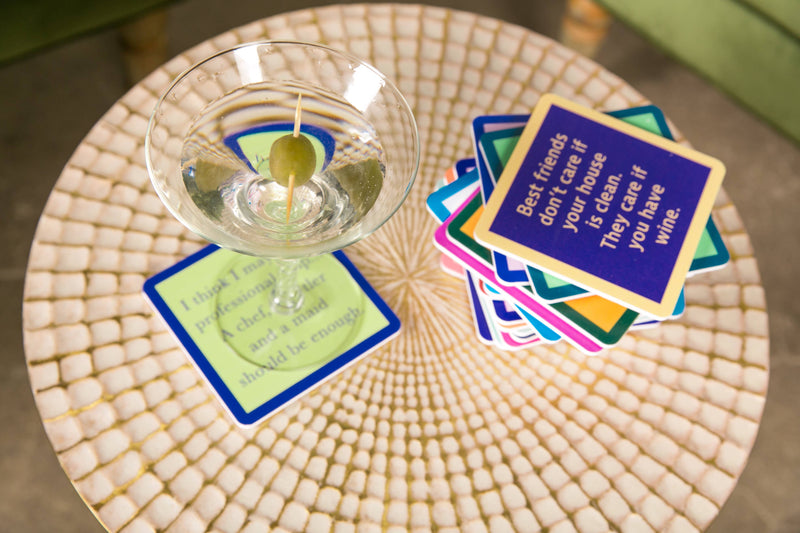 Drinks on Me coaster with a funny quote, accompanies a martini glass on a mosaic table, emphasizing playful home bar decor for Athens Cooks.