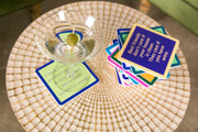 Round side table with Drinks on Me - COASTER Nervous featuring witty text beside a martini glass and stack of colorful coasters, ideal for home bar decor.
