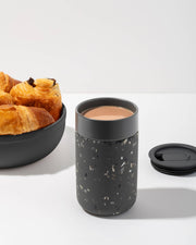 W&P - Ceramic Reusable Coffee Mug 16oz: Charcoal, featuring a grippable silicone sleeve and splash-resistant lid, pictured beside a coffee cup and croissants.