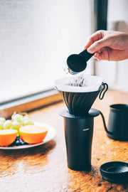 HARIO USA - V60 Metal Dripper: Silver in use, with coffee grounds being added, atop a travel mug, surrounded by a fruit plate and gooseneck kettle.