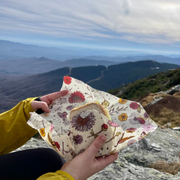 Person in a yellow jacket holding a sandwich wrapped in Bee's Wrap - Cut-to-Size Roll - Meadow Magic, atop a mountain overlook, showcasing sustainable food wrapping.
