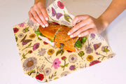 Bee's Wrap - Sandwich Wrap - Meadow Magic being folded around a deli sandwich on a countertop, showcasing its floral design and secure wooden bee button closure.
