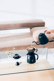 Minimalist pour-over setup featuring HARIO USA - V60 Metal Dripper: Silver, with gooseneck kettle, coffee server, and accessories on a glass table.