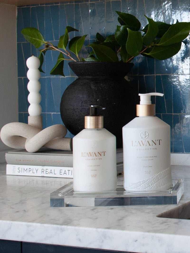 L'AVANT Collective - Fresh Linen Hand Lotion in a glass dispenser on a marble counter, accompanied by a black vase with green leaves.