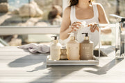 L'AVANT Collective Hand Lotion Ambre Santal on marble tray, alongside soaps and brush, in a sunlit kitchen, with a person washing dishes in the background.