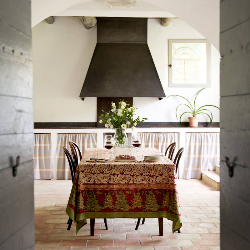 Couleur Nature - Noel Red & Green tablecloth adorns a cozy dining nook with bent-wood chairs, wine, and a floral centerpiece, evoking festive, French-inspired charm.