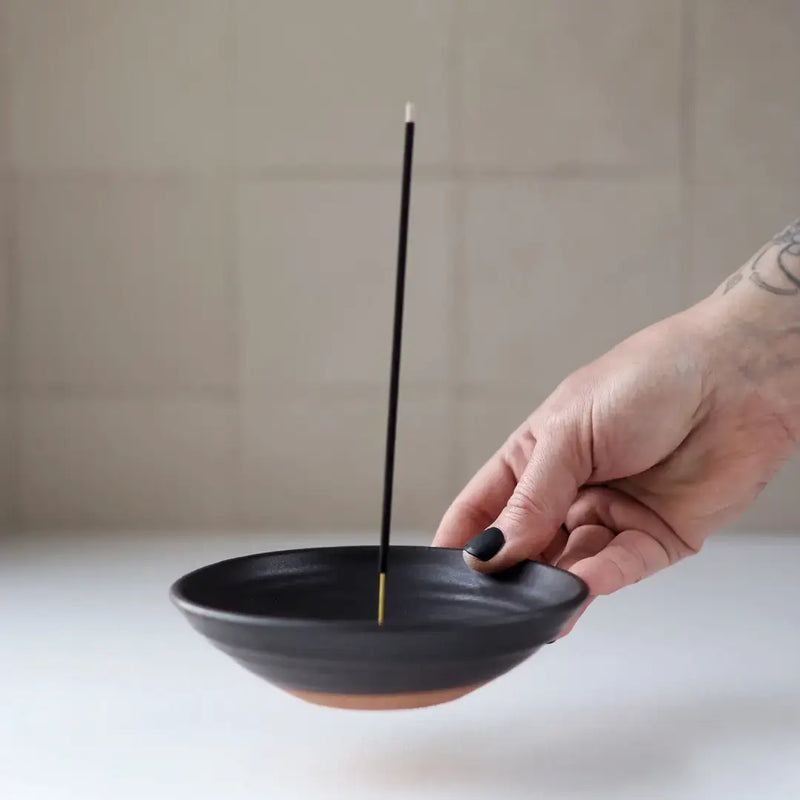 Hand holding a Gravesco Pottery - Incense Burner in black ceramic, showcasing its terracotta base and upright incense stick, ideal for catching ash elegantly.