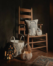 Coral & Tusk - Scaredy Black Cat Pillow sits on a rustic chair, surrounded by a fabric pumpkin basket and decorative containers, creating a cozy Halloween-inspired vignette.