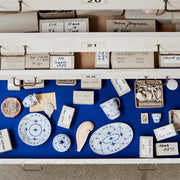 Royal Copenhagen | Blue Fluted Plain Cups, Set of 2, displayed in a museum-like drawer with porcelain shards and artifacts, highlighting craftsmanship and classic design.