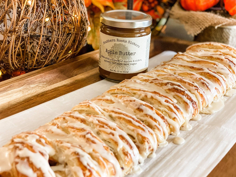 Southern Roots Sisters Apple Butter jar beside a braided pastry with icing, set on a wooden board amidst autumn decorations, showcasing a cozy, fall-themed ambiance.