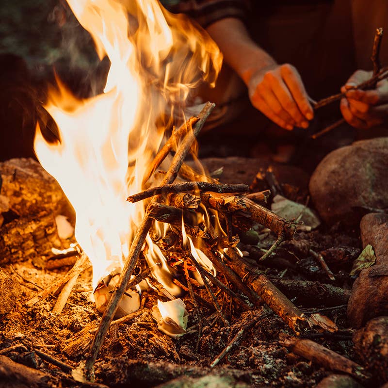 Hands arranging sticks into a campfire with Bee's Wrap - ReKindle Fire Starters™, showcasing sustainable, lightweight strips perfect for camping and reducing waste.