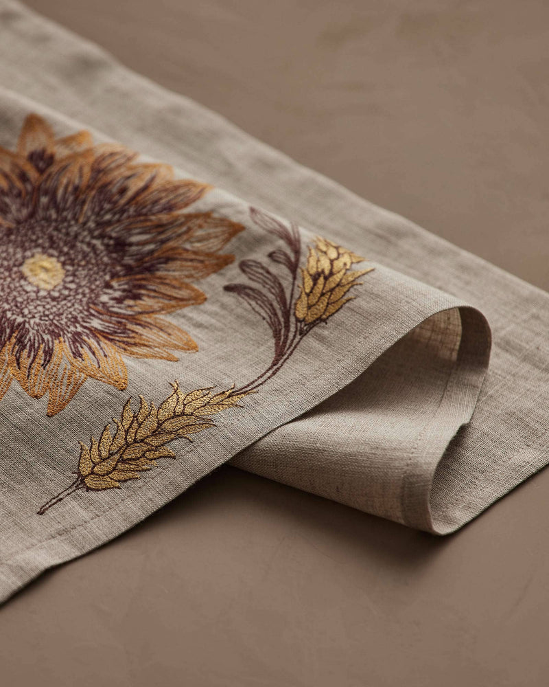 Coral & Tusk - Sunflower and Wheat Dinner Napkin: Close-up of embroidered sunflower and wheat motif on folded linen napkin, ideal for autumnal table settings.