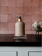 L'AVANT Collective - High Performing Hand Soap - Blushed Bergamot, in a blush pink glass bottle on a counter, near flowers.
