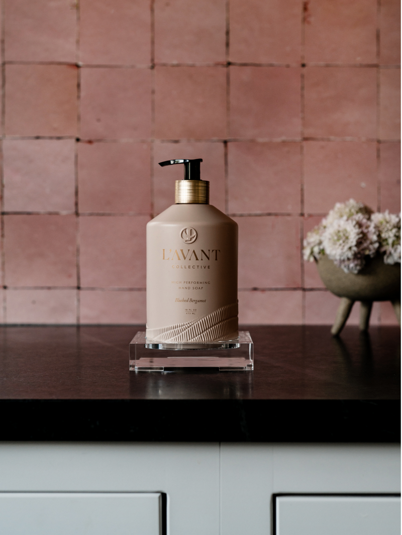 L'AVANT Collective - High Performing Hand Soap - Blushed Bergamot, in a blush pink glass bottle on a counter, near flowers.