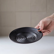 Gravesco Pottery - Garlic Grater Plate in Black held up, showcasing its concentric spiral texture for efficient garlic preparation and serving, ideal for enhancing kitchen rituals.