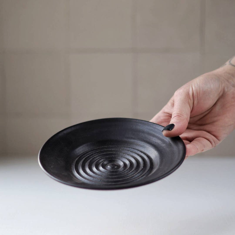 Gravesco Pottery - Garlic Grater Plate in Black held up, showcasing its concentric spiral texture for efficient garlic preparation and serving, ideal for enhancing kitchen rituals.
