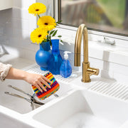 Hand scrubbing saucepan with Euroscrubby in a bright kitchen sink area, featuring a double-basin sink, brass faucet, and blue accessories on the windowsill.