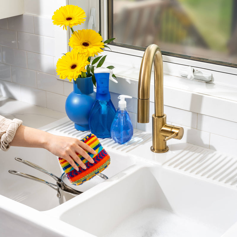 Hand scrubbing saucepan with Euroscrubby in a bright kitchen sink area, featuring a double-basin sink, brass faucet, and blue accessories on the windowsill.