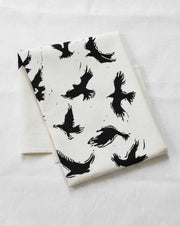 Hearth and Harrow Organic Cotton Flying Crows Tea Towel, featuring black raven silhouettes on off-white fabric, hand-printed with eco-friendly ink and a twill hanging loop.