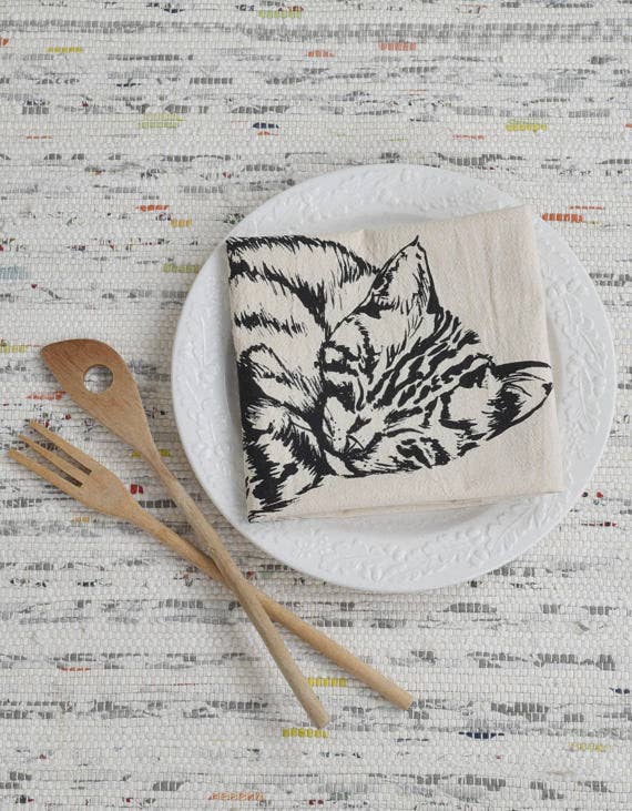 Hearth and Harrow - Organic Cotton Sleeping Cat Tea Towel in Black displayed on a ceramic plate, accompanied by wooden utensils on a woven background.