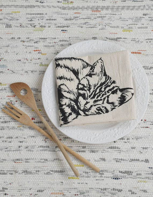 Hearth and Harrow - Organic Cotton Sleeping Cat Tea Towel in Black displayed on a ceramic plate, accompanied by wooden utensils on a woven background.