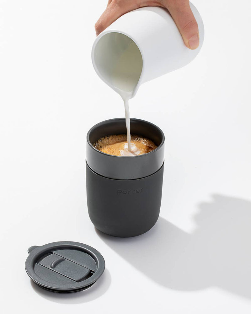 Person pouring milk into W&P - Reusable Ceramic Mug: Terrazzo Charcoal, featuring a splash-resistant lid and silicone sleeve, perfect for sustainable home brewing.