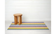 Chilewich | Pop Stripe Shag Mat - 24 x 36 displayed on a floor with a wooden stool, highlighting its durable woven textile and aesthetic appeal.