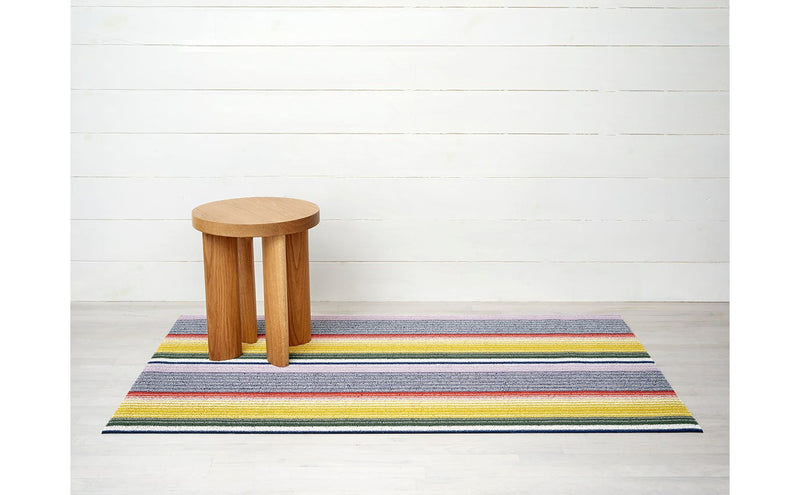 Chilewich | Pop Stripe Shag Mat - 24 x 36 displayed on a floor with a wooden stool, highlighting its durable woven textile and aesthetic appeal.
