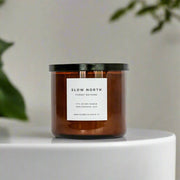 Slow North - Forest Bathing 3-Wick Non-Toxic Soy Candle with essential oils in a brown glass jar, black lid, surrounded by soft-focus greenery, invoking a serene, earthy ambiance.