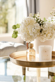 L'AVANT Collective Fresh Linen Room Spray 100 ml bottle and white candle on a gold tray, adding elegant fragrance to your home.