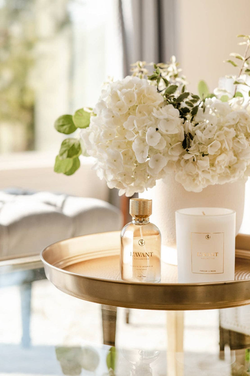 L'AVANT Collective Fresh Linen Room Spray 100 ml bottle and white candle on a gold tray, adding elegant fragrance to your home.