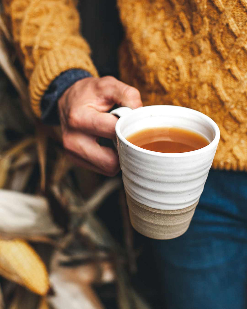 Close-up of a person holding a Farmhouse Pottery - Tall Silo Mug filled with tea, highlighting its handcrafted, two-tone design. Perfect for sipping large in style.